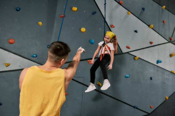 Male instructor holding rope, talking to a girl in safety equipment and harness while she is training on the artificial climbing wall - Foto, immagini