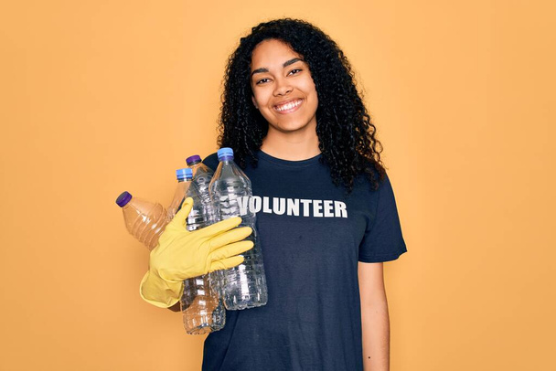 Jeune femme afro-américaine faisant du bénévolat recyclage tenant des bouteilles en plastique avec un visage heureux debout et souriant avec un sourire confiant montrant les dents - Photo, image