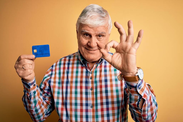 Senior handsome hoary customer man holding credit card to finance payment doing ok sign with fingers, excellent symbol - Valokuva, kuva