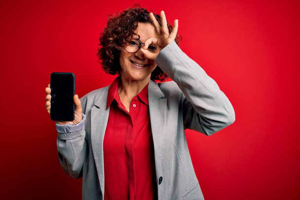 Middle age curly woman holding smartphone showing screen over isolated red background with happy face smiling doing ok sign with hand on eye looking through fingers - Fotó, kép