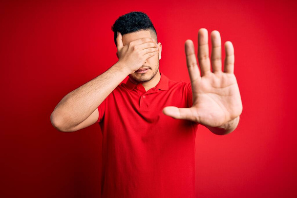 Jonge knappe man draagt rode casual polo staan over geïsoleerde achtergrond bedekken ogen met handen en doen stop gebaar met droevige en angst uitdrukking. Beschaamd en negatief concept. - Foto, afbeelding