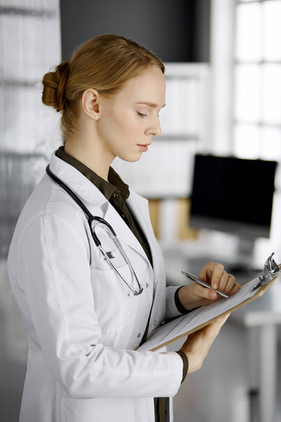 Friendly smiling female doctor using clipboard in clinic. Portrait of friendly physician woman at work. Medical service in hospital. Medicine concept - Foto, imagen