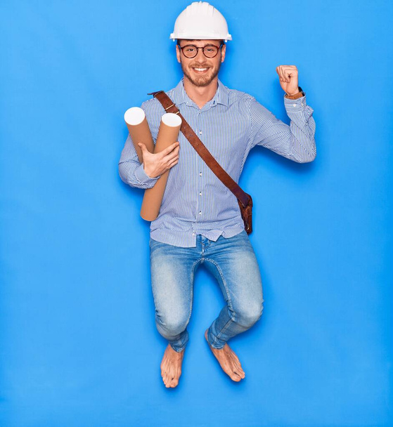Young handsome architect man wearing leather bag and hardhat smiling happy. Jumping with smile on face holding blueprints celebrating with fist raised over isolated blue background - 写真・画像