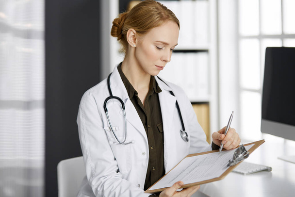 Cheerful smiling female doctor using clipboard in clinic. Portrait of friendly physician woman at work place. Perfect medical service in hospital. Medicine concept - Foto, imagen