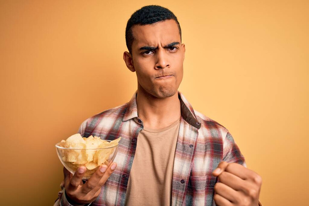 Молодой красивый африканский американец держит миску чипсов картофель на желтом фоне раздраженный и разочарованный кричать от гнева, сумасшедший и кричать с поднятой рукой, гнев концепции
 - Фото, изображение