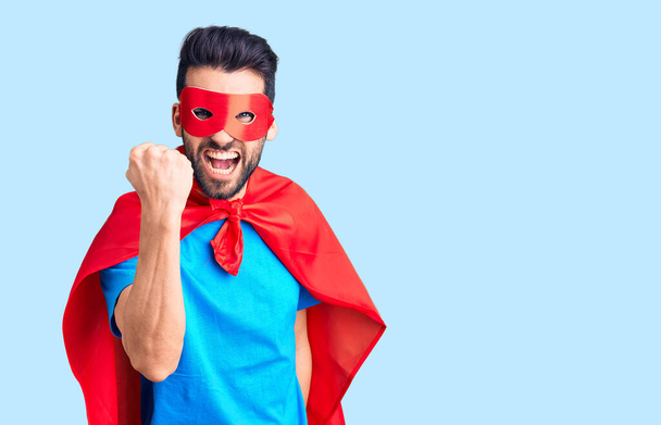 Young handsome man with beard wearing super hero costume angry and mad raising fist frustrated and furious while shouting with anger. rage and aggressive concept.  - Photo, Image