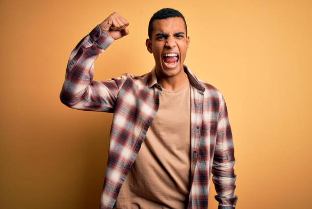 Junger gutaussehender afrikanisch-amerikanischer Mann in lässigem Hemd, der vor gelbem Hintergrund steht und wütend und wütend die Faust erhebt, während er vor Wut schreit. Wut und aggressives Konzept. - Foto, Bild