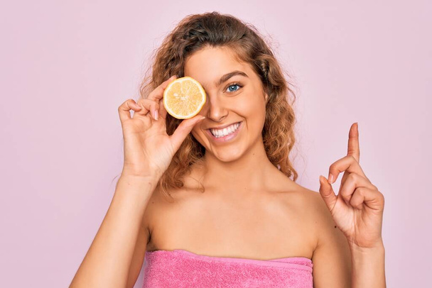 Beautiful woman with blue eyes wearing towel shower after bath holding lemon fruit over eye surprised with an idea or question pointing finger with happy face, number one - Fotografie, Obrázek