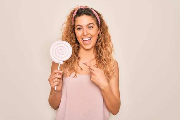Giovane bella donna bionda con gli occhi blu mangiare caramelle dolci lecca-lecca su sfondo rosa molto felice indicando con mano e dito - Foto, immagini
