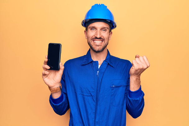 Mladý pohledný dělník v uniformě a klobouku drží smartphone zobrazující obrazovku křičí hrdě, slaví vítězství a úspěch velmi vzrušený se zvednutou paží - Fotografie, Obrázek