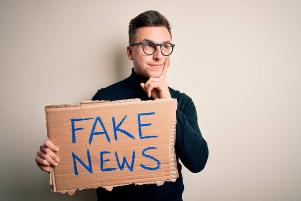 Junger gutaussehender kaukasischer Mann, der Protest-Karton für Desinformation und Fake News in der Hand hält, ernstes Gesicht über Frage nachdenkend, sehr verworrene Idee - Foto, Bild