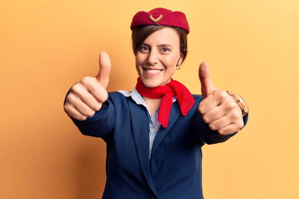 Young beautiful woman wearing stewardess uniform approving doing positive gesture with hand, thumbs up smiling and happy for success. winner gesture.  - Foto, Bild