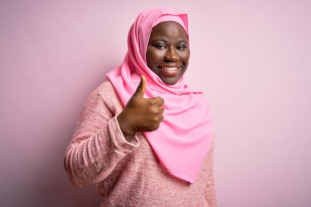 Νεαρή Αφροαμερικάνα συν μέγεθος γυναίκα φορώντας μουσουλμανική μαντίλα πάνω από απομονωμένο ροζ φόντο κάνει ευτυχισμένη αντίχειρες επάνω χειρονομία με το χέρι. Έγκριση έκφρασης κοιτάζοντας την κάμερα που δείχνει επιτυχία. - Φωτογραφία, εικόνα