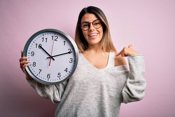 Jeune belle femme brune portant des lunettes faisant le compte à rebours en utilisant une grande horloge avec visage surprise pointant du doigt vers lui-même - Photo, image