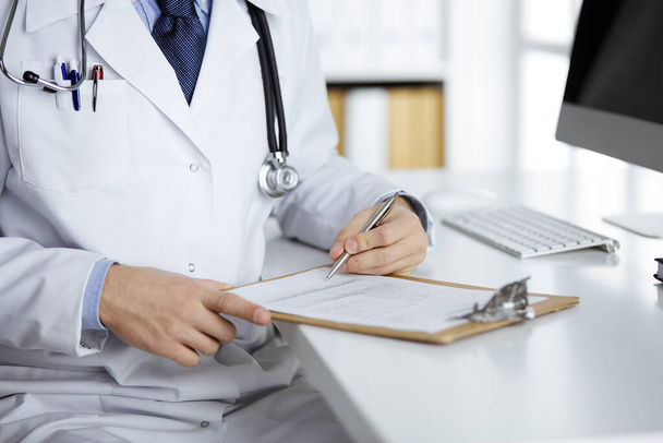 Friendly red-bearded doctor sitting and writing with clipboard in clinic, close-up. Medicine concept - Foto, imagen