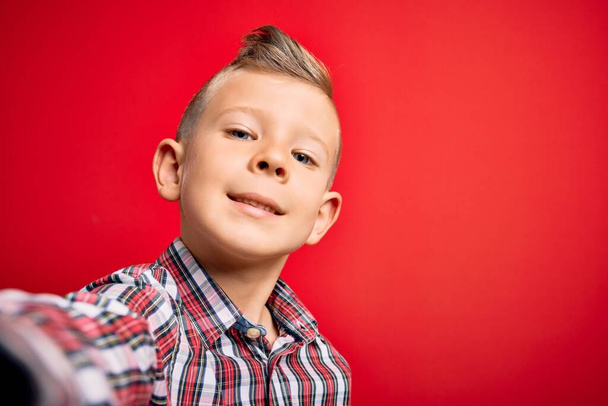 Close up de criança caucasiana jovem com olhos azuis tirando uma foto selfie sobre fundo vermelho com um rosto feliz de pé e sorrindo com um sorriso confiante mostrando dentes
 - Foto, Imagem