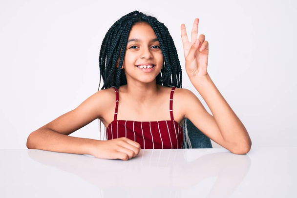 Молодая африканская девочка с косичками в повседневной одежде сидит на столе и показывает пальцами номер два улыбаясь уверенно и счастливо.  - Фото, изображение