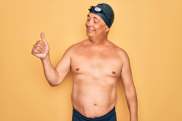 Middle age senior grey-haired swimmer man wearing swimsuit, cap and goggles Looking proud, smiling doing thumbs up gesture to the side - Fotoğraf, Görsel