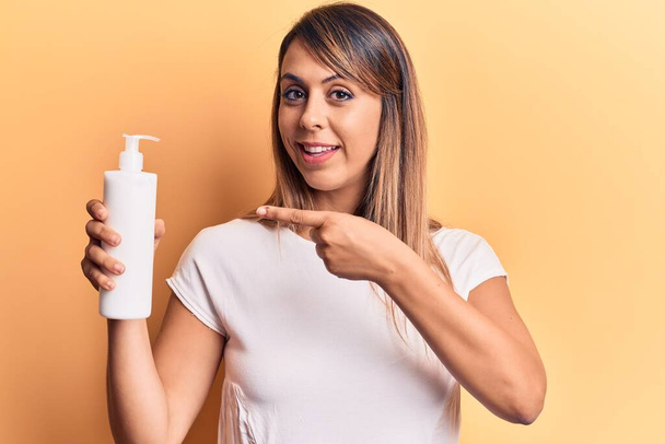 Young beautiful woman holding bottle of cream lotion smiling happy pointing with hand and finger  - Фото, зображення