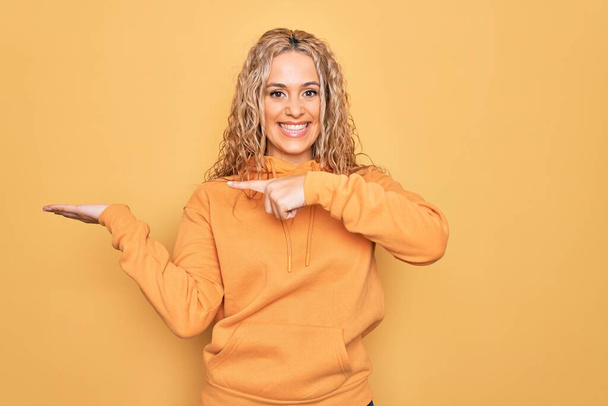 Młoda piękna blondynka sportowa kobieta w luźnej bluzie na żółtym tle zdumiona i uśmiechnięta do aparatu, prezentująca się z ręką i wskazująca palcem. - Zdjęcie, obraz