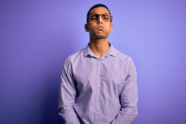 Bel homme afro-américain portant une chemise rayée et des lunettes sur fond violet joues gonflées avec un visage drôle. Bouche gonflée avec de l'air, expression folle. - Photo, image