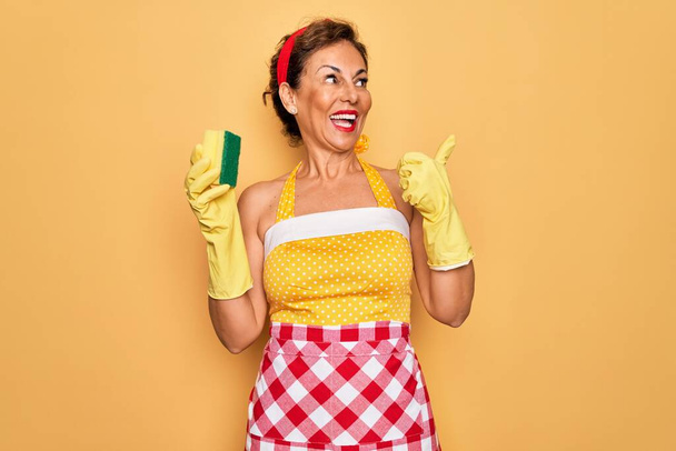 Orta yaşlı yaşlı ev hanımı, 50 'li yılların tarzı retro elbiseler giyen, daha temiz fırça kullanan ve baş parmağıyla gülümseyen bir kadın tasması takıyor. - Fotoğraf, Görsel