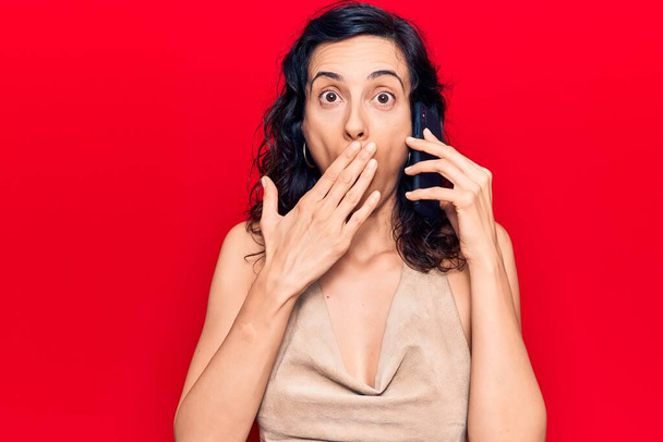 Junge schöne hispanische Frau im Gespräch mit dem Smartphone, das Mund mit der Hand bedeckt, schockiert und Angst vor Fehlern. Überraschte Miene  - Foto, Bild
