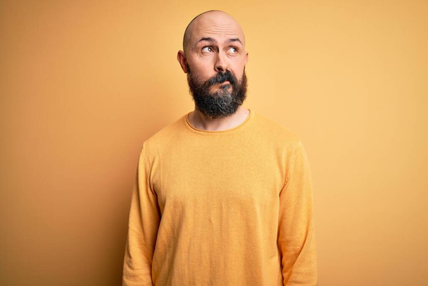 Красивый лысый мужчина с бородой в обычном свитере стоит на жёлтом фоне улыбается, смотрит в сторону и думает:
. - Фото, изображение