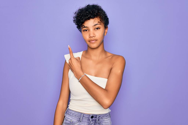 jong mooi Afrikaans amerikaans afro vrouw dragen casual t-shirt over paarse achtergrond wijzend met de hand vinger naar de zijkant tonen reclame, ernstige en rustige gezicht - Foto, afbeelding