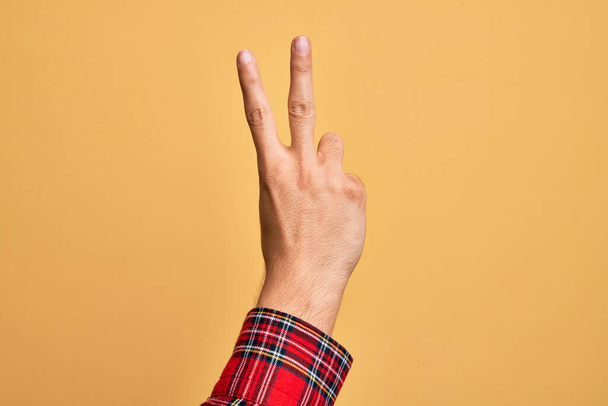 Mano di giovane caucasico che mostra le dita su uno sfondo giallo isolato contando il numero 2 mostrando due dita, gesticolando vittoria e simbolo del vincitore - Foto, immagini