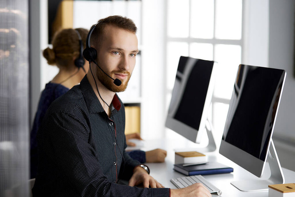 Red-bearded businessman talking by headset near his female colleague while sitting in modern office. Diverse people group in call center. Telemarketing and customer service - Foto, immagini