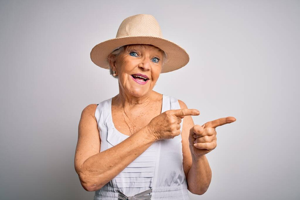 Senior beautiful grey-haired woman on vacation wearing casual summer dress and hat smiling and looking at the camera pointing with two hands and fingers to the side. - Photo, image