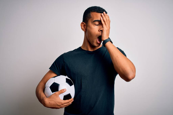 Нахабний афро-американець, який грає у футбол, тримаючи м'яч на білому фоні, і втомлений, закриваючи половину обличчя, очі і рот рукою. Біль завдає болю. - Фото, зображення