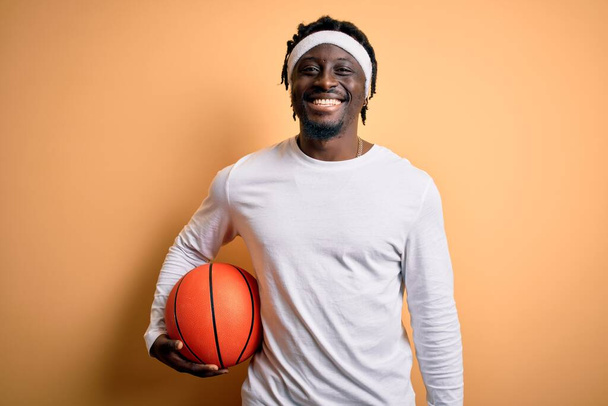 若いアフリカ系アメリカ人のスポーツマンが黄色の背景にバスケットボールボールを保持し、幸せな顔をして笑顔で歯を示す自信を持って笑顔で - 写真・画像