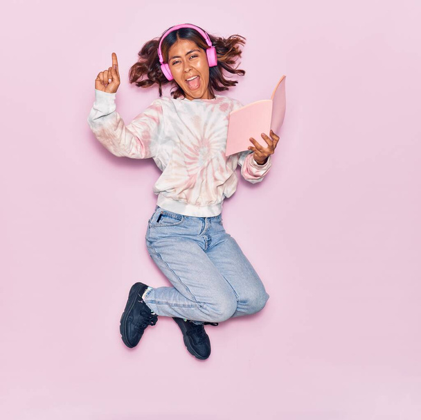 Young beautiful latin woman listening to music using headphones smiling happy. Jumping with smile on face reading book over isolated pink background - 写真・画像
