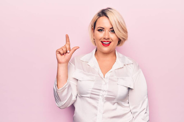 Young beautiful blonde plus size woman wearing elegant shirt over isolated pink background smiling with an idea or question pointing finger up with happy face, number one - 写真・画像