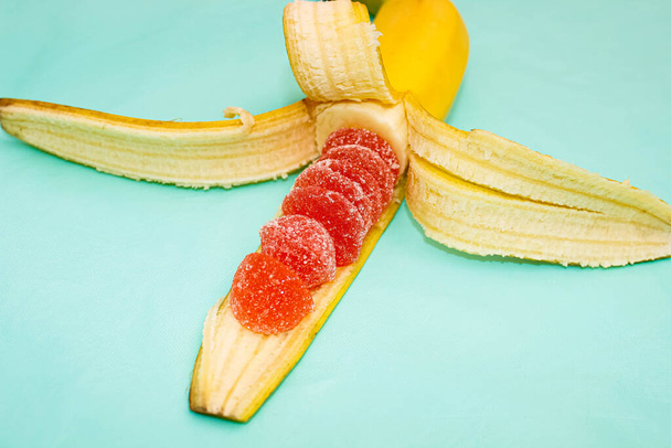 Полуочищенный банан, внутри которого вместо фруктово-красного мармелада в сахаре. Селективный фокус, бирюзовый фон
. - Фото, изображение