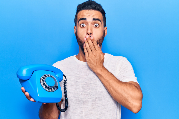 Mladý hispánec držící starodávný telefon za ústa rukou, šokovaný a vystrašený z omylu. překvapený výraz  - Fotografie, Obrázek