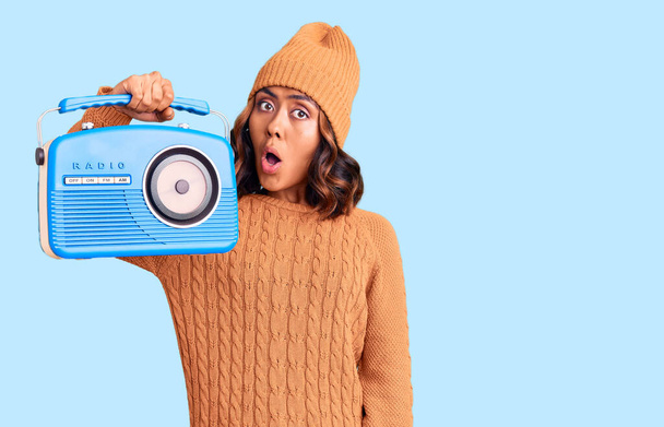 Молодая красивая смешанная раса женщина держит винтажное радио испуганы и поражены с открытым ртом для удивления, неверие лицо  - Фото, изображение
