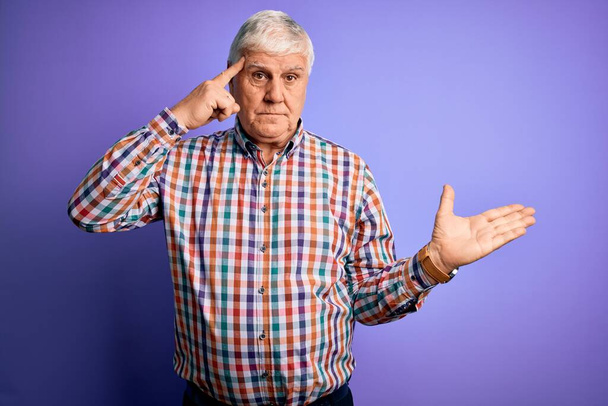 Starší pohledný chraptivý muž v nenucené barevné košili přes izolované purpurové pozadí zmatený a otrávený s otevřenou dlaní ukazující kopírovací prostor a ukazující prstem na čelo. Přemýšlej o tom.. - Fotografie, Obrázek