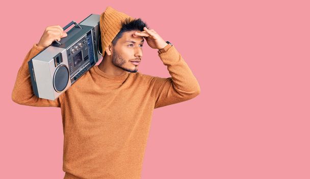 Handsome latin american young man holding boombox, listening to music very happy and smiling looking far away with hand over head. searching concept.  - Photo, Image