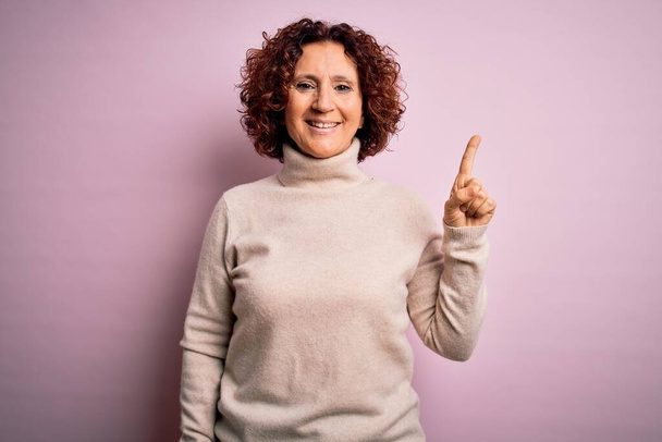 Mujer de mediana edad hermosa cabello rizado con suéter de cuello alto casual sobre fondo rosa con una gran sonrisa en la cara, señalando con el dedo de la mano al lado mirando a la cámara
. - Foto, Imagen