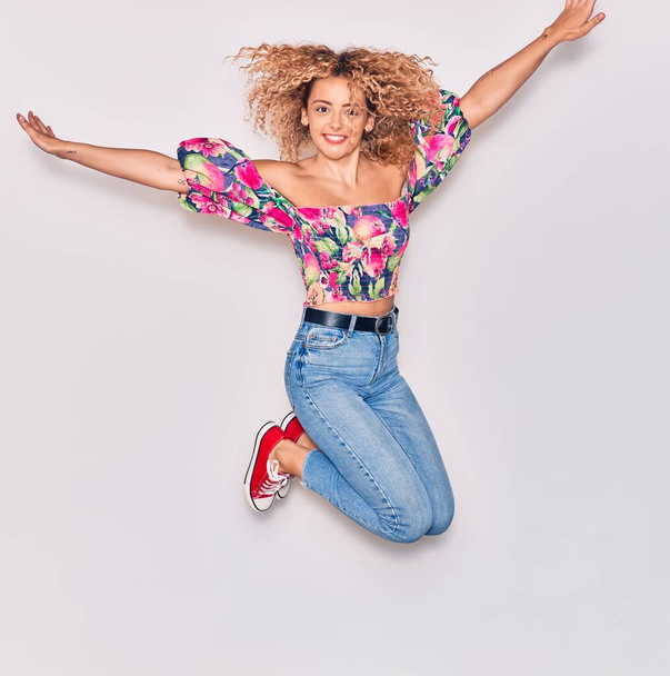 Young beautiful curly girl wearing casual clothes smiling happy. Jumping with smile on face and arms opened over isolated white background. - Valokuva, kuva