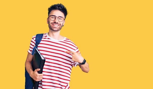 Jeune homme hispanique portant un sac à dos étudiant et des lunettes tenant le cartable pointant du doigt un sourire heureux et fier  - Photo, image