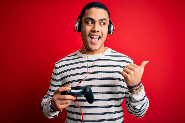 若いブラジルのゲーマーの男はジョイスティックとヘッドフォンを使用してビデオゲームをプレイし、幸せな顔の笑顔で親指で側まで示す - 写真・画像