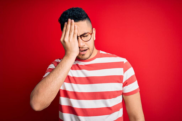 Young handsome man wearing casual striped t-shirt and glasses over isolated red background Yawning tired covering half face, eye and mouth with hand. Face hurts in pain. - Fotografie, Obrázek