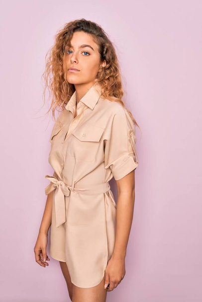 Young beautiful blonde woman with curly hair wearing casual dress standing over isolated pink background - Zdjęcie, obraz