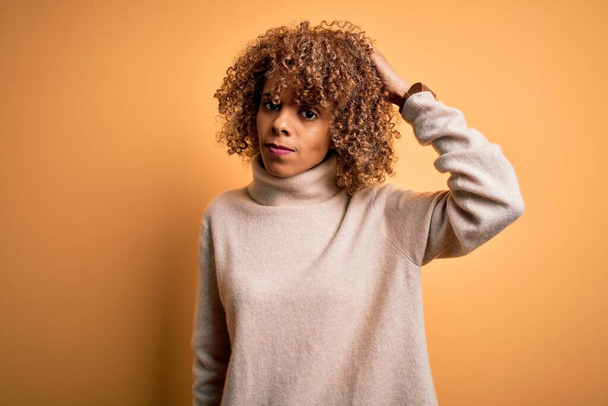 Young beautiful african american woman wearing turtleneck sweater over yellow background confuse and wondering about question. Uncertain with doubt, thinking with hand on head. Pensive concept. - Photo, Image