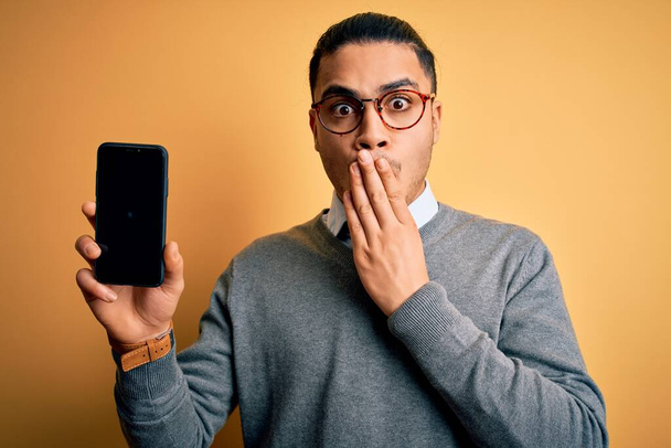 Młody brazylijski biznesmen w okularach trzymający smartfona pokazujący pokrywę ekranu ustami z ręką zaszokowany wstydem za pomyłkę, wyrażaniem strachu, przerażony w ciszy, sekretny koncept - Zdjęcie, obraz