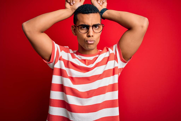 Young handsome african american man wearing casual striped t-shirt and glasses doing funny gesture with finger over head as bull horns - Photo, Image